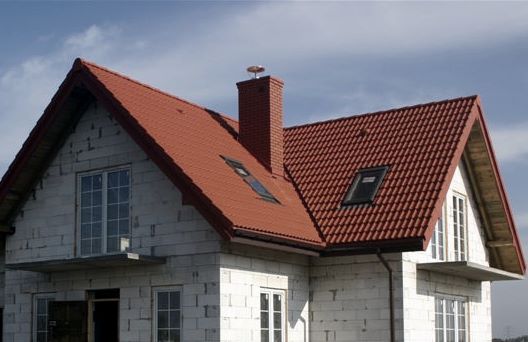 Dom w trakcie budowy z czerwoną dachówką ceramiczną, widoczne okna dachowe i komin z anteną, ściany z białego bloczka.